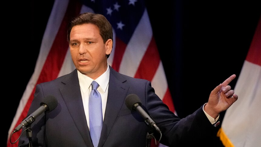 A middle-aged white man with brown hair wearing a suit speaks in front of an American flag.