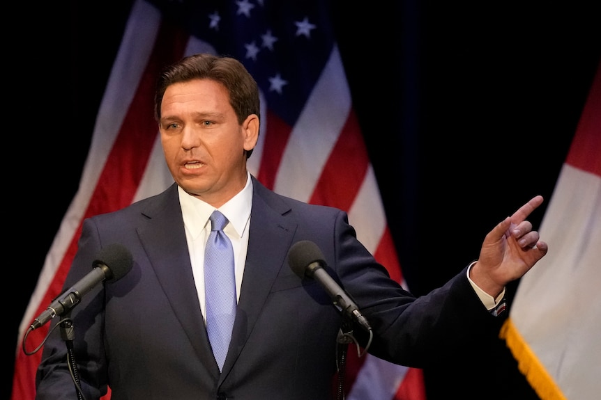 A middle-aged white man with brown hair wearing a suit speaks in front of an American flag.