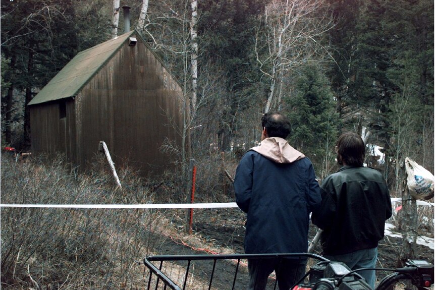 Two men are seen looking at a small cabin in the woods