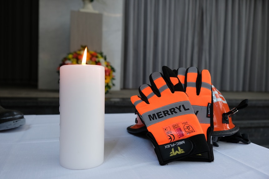 a pair of hi vis orange coloured gloves inscribed with the name Merryl sits on a white altar next to a white candle which is lit