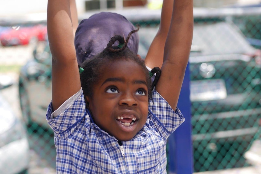 A young girl hands from bars in the school yard with her arms outstretched and another student behind her.