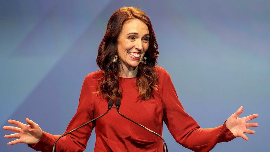 Jacinda Ardern smiles as she stands in front of a blue background and holds her hands out on either side of her.