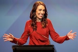 Jacinda Ardern smiles as she stands in front of a blue background and holds her hands out on either side of her.