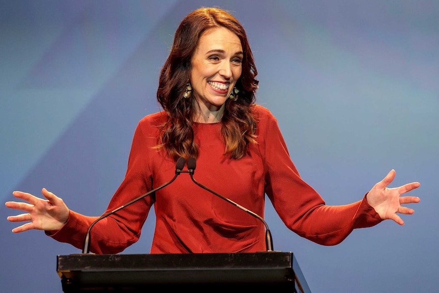 Jacinda Ardern smiles as she stands in front of a blue background and holds her hands out on either side of her.