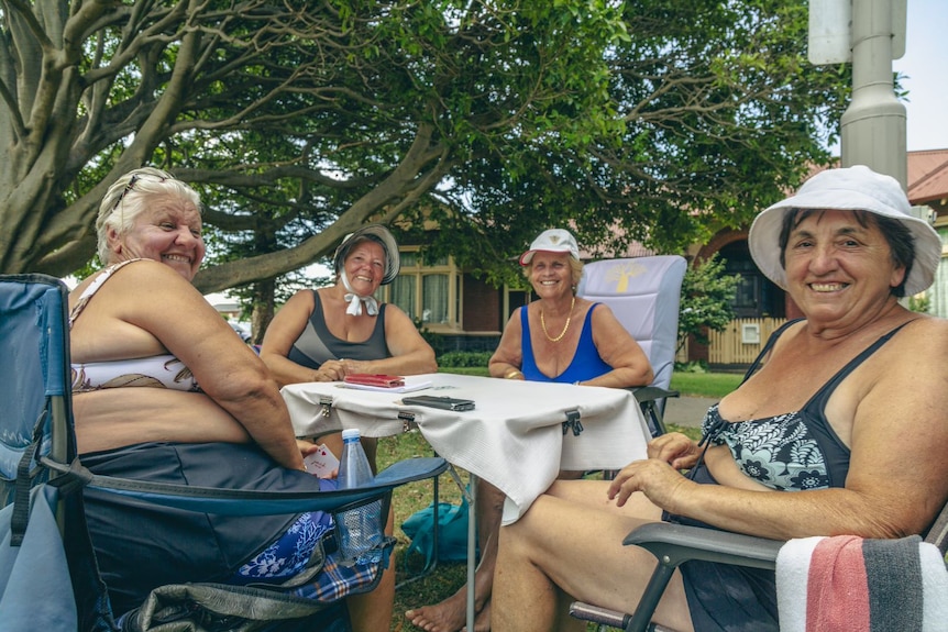 Four women sit at a table in a park, they wear bathers and play cards