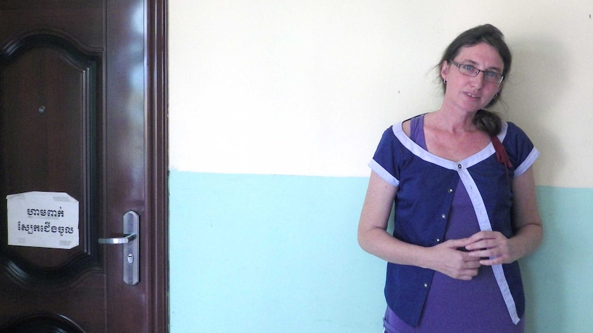 A woman in a purple top and wearing glasses stands next to a white wall