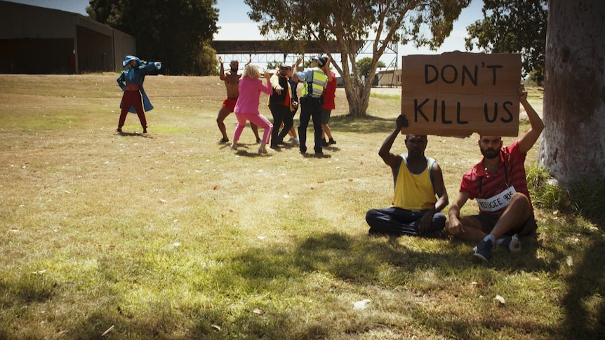 Two men sit and hold a sign saying 'don't kill us' and in the background a man in a Captain Cook costume points at dancers