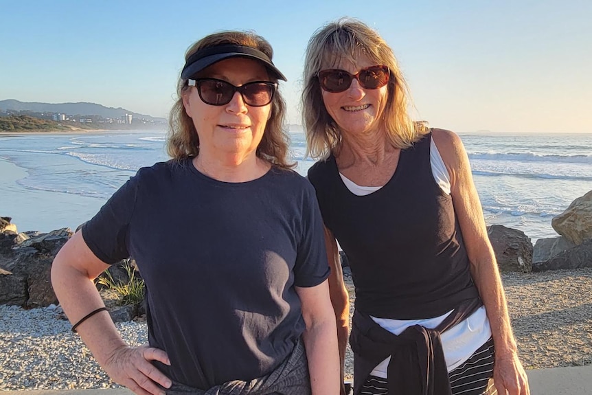 Two women wearing t shirts and sunglasses, beach in the background