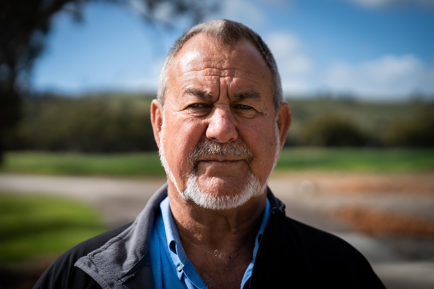 Portrait of a man with a weathered face, short hair, neat beard, standing on a sunny property lined with trees.