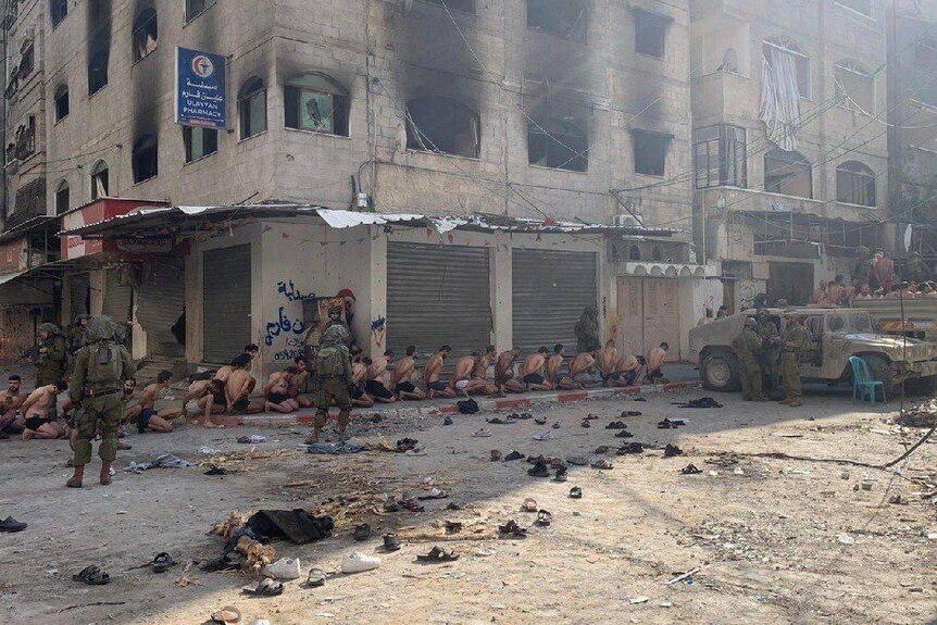 A damaged street with military vehicles on the right, soldiers and a queue of men bound, kneeled and stripped down