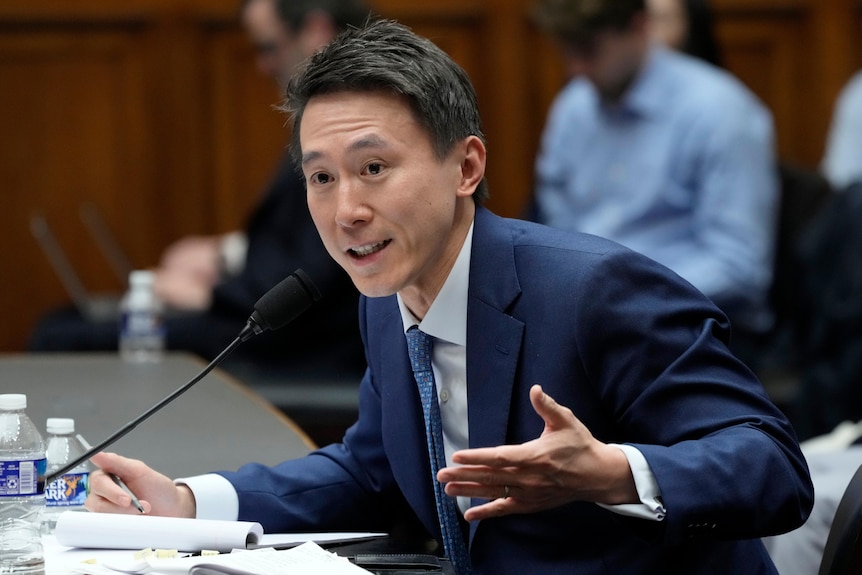 A man wearing a blue suit is sitting at a table behind a microphone. He gestures with his left hand as he speaks.