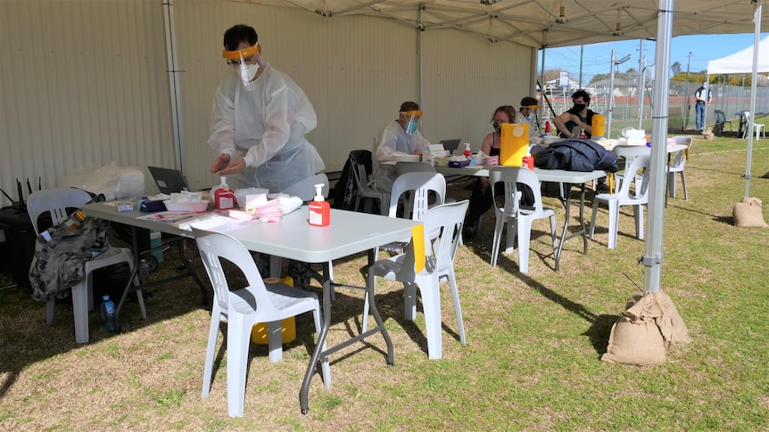 A medical professional stands behind a table under a white tent, preparing a vaccination needle