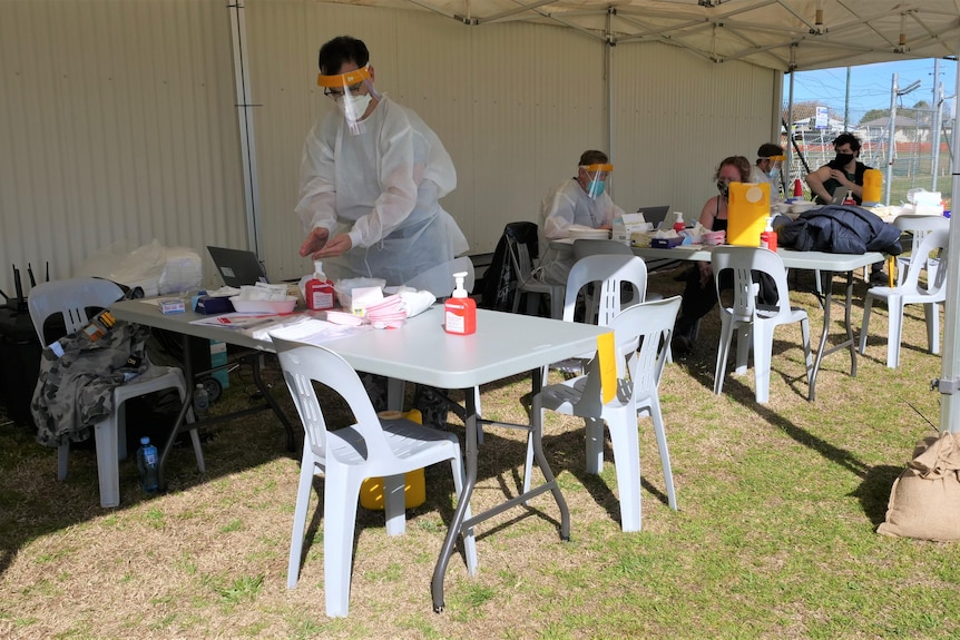 A medical professional stands behind a table under a white tent, preparing a vaccination needle.
