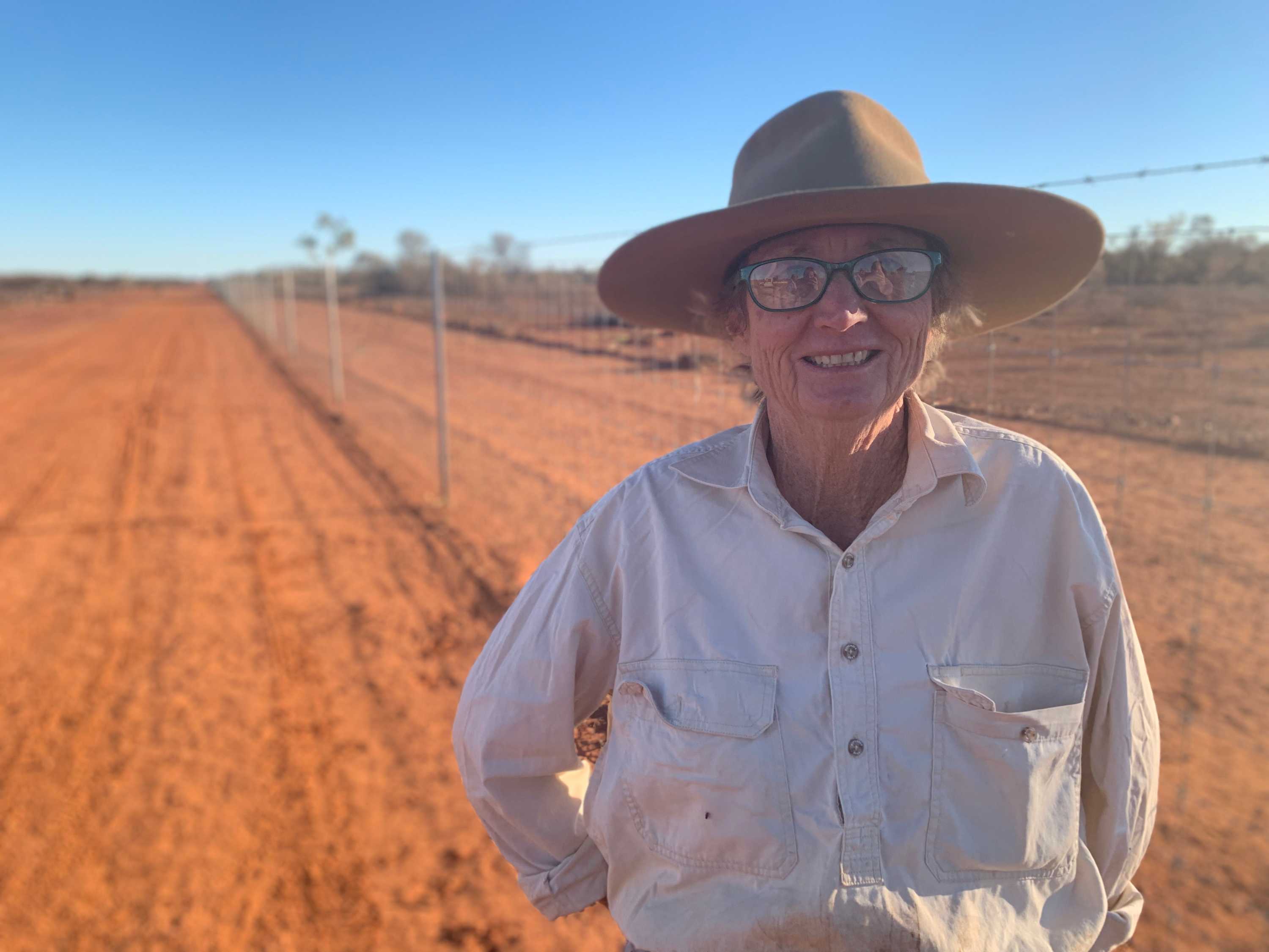 A woman stands by a long tall fence in the red dust of western Queensland