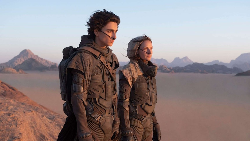 Two actors dressed in futuristic armour in the foreground of a vast desert, staring into the distance.