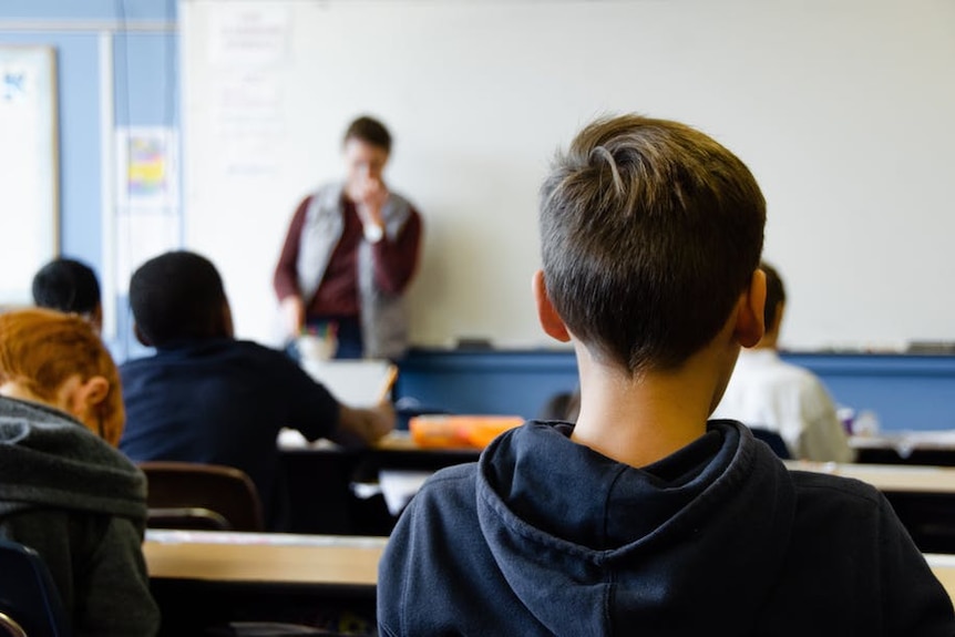 A boy sits in class looking forward to a teacher.