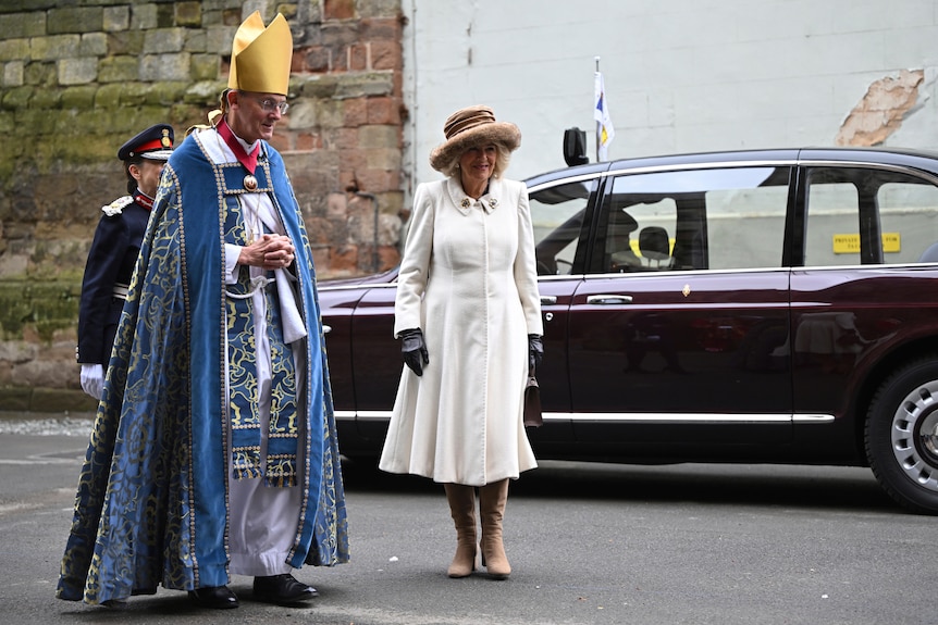 Camilla is wearing a hat and long white dress. She's standing next to a bishop.