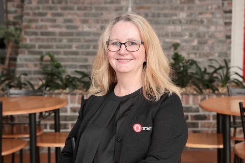 A blonde-haired woman wearing glasses and a black jacket smiles at the camera sitting in a cafe.