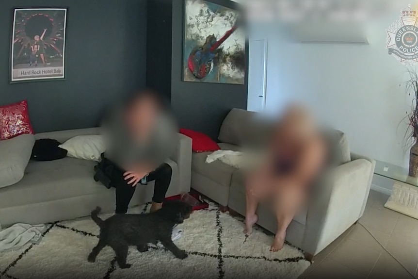 A blurred image of a police officer sitting next to an accused on a couch inside a home. 
