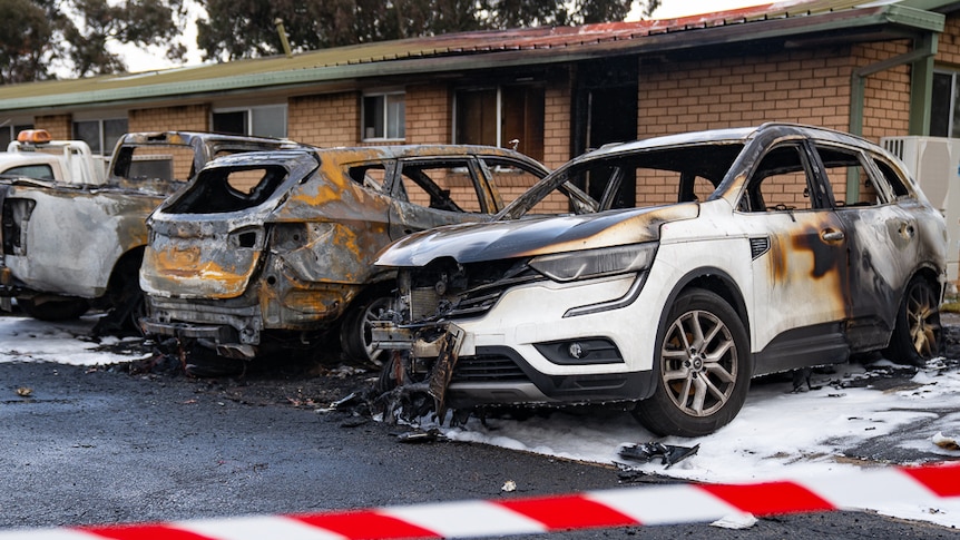 Three burnt out cars in front of a fire damaged building.