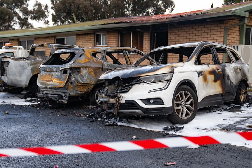 Three burnt out cars in front of a fire damaged building.