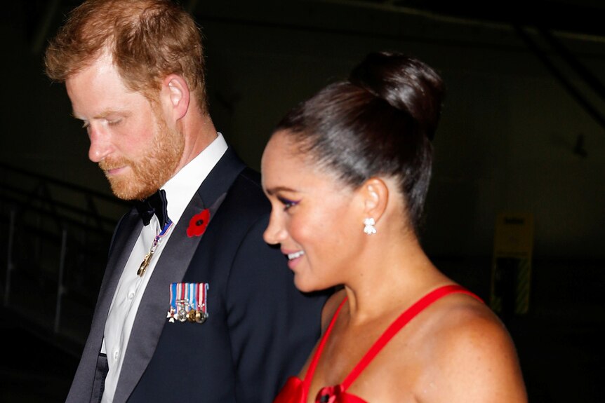 Prince Harry dressed in a suit and Meghan Markle, wearing a red dress walk at an event