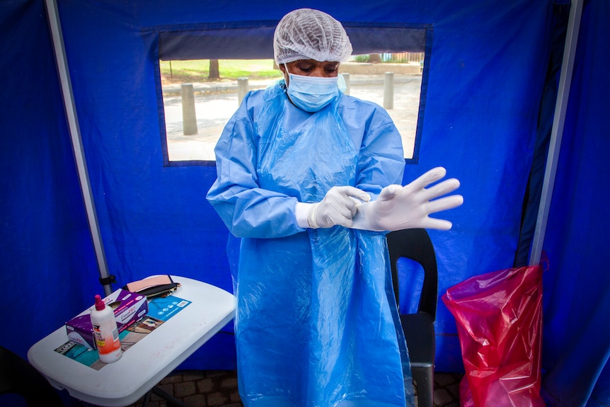 A man in blue scrubs and a face mask adjusts his gloves in a medical tent 
