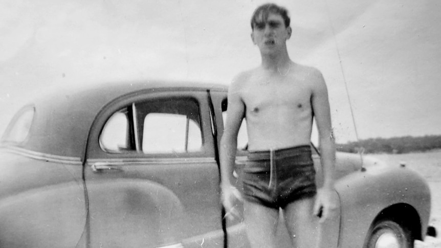 A black and white photo of a man in bathers in the 1960s standing next to an old car