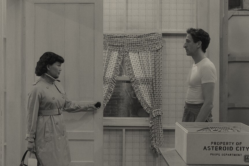 A black and white shot of an Asian woman in a trench coat & a white man with dark hair in a white t-shirt standing in a doorway