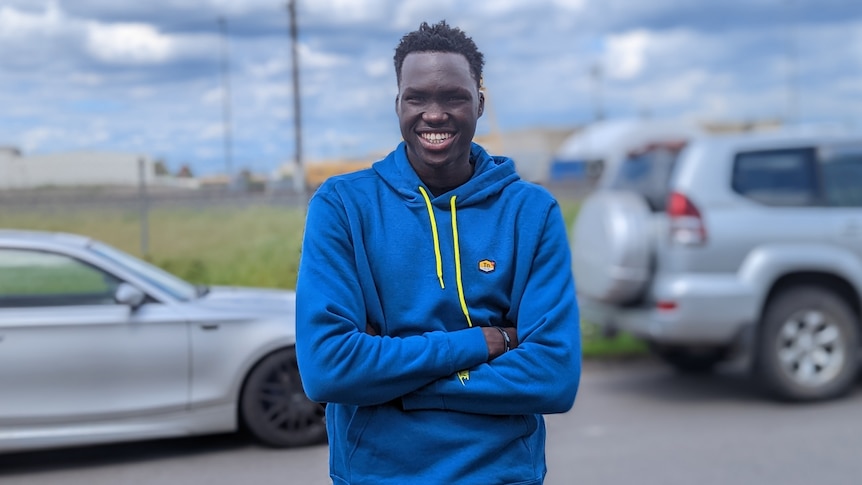A smiling young man in a blue hoodie stands in front of a road.