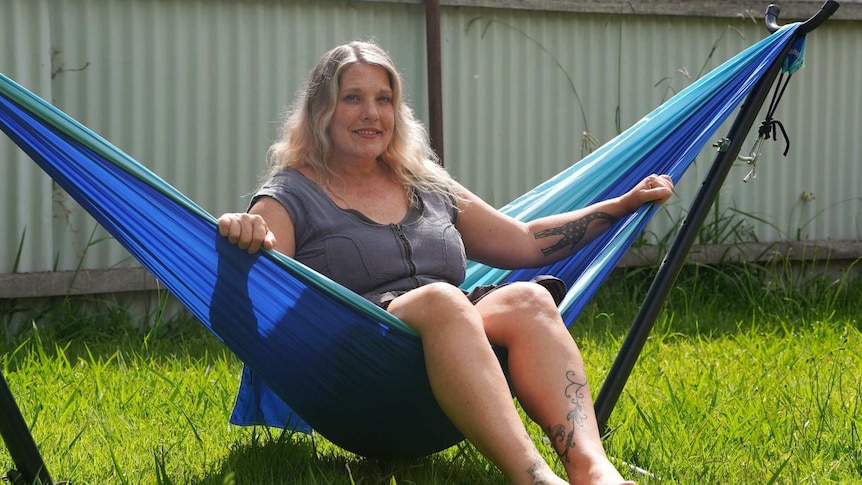 A blonde haired woman reclines in a bright blue  hammock while the sun shines down onto green grass