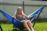 A blonde haired woman reclines in a bright blue  hammock while the sun shines down onto green grass