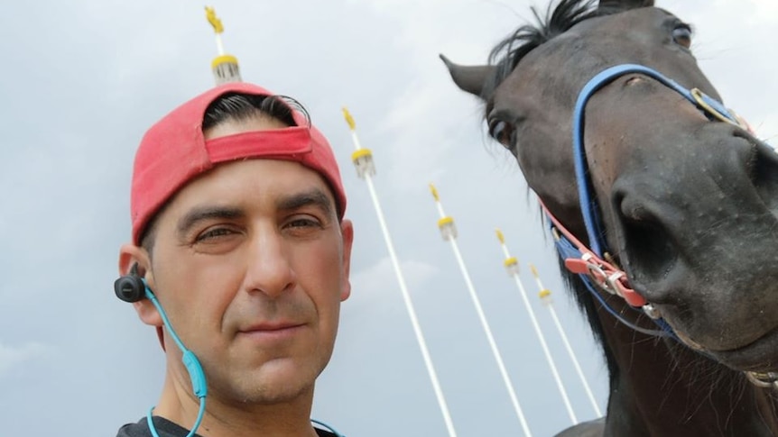 A selfie of a man with a backwards red cap and headphones next to a dark horse in bridle.