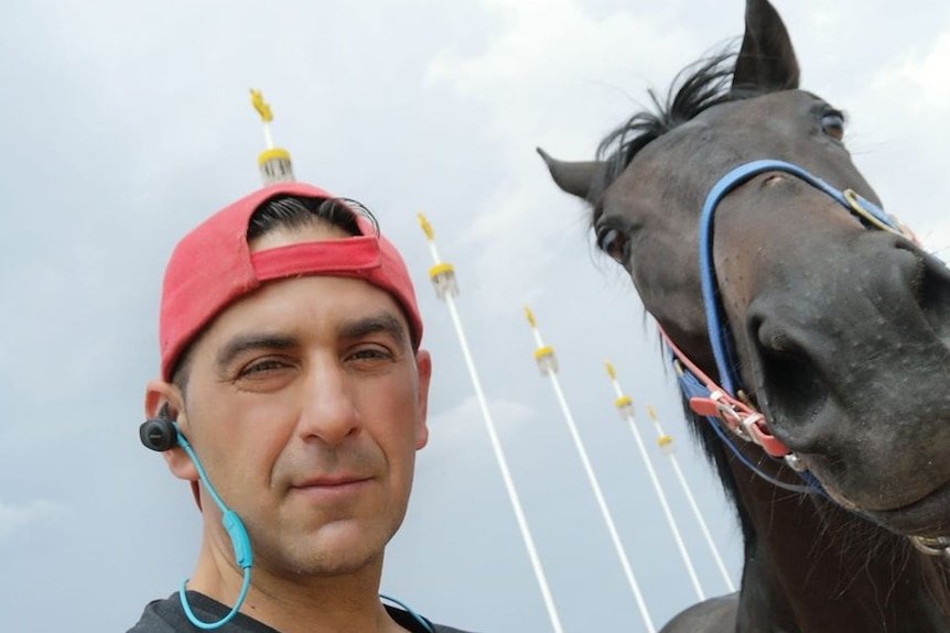 A selfie of a man with a backwards red cap and headphones next to a dark horse in bridle.