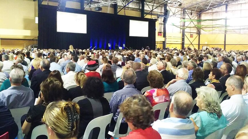 Hundreds of people face two screens and podium in a Gatton sports hall for the funeral of Steve Jones