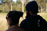 A man and woman stand silhouetted looking out at sunny trees and grass, wearing beanie and sunglasses.