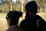 A man and woman stand silhouetted looking out at sunny trees and grass, wearing beanie and sunglasses.