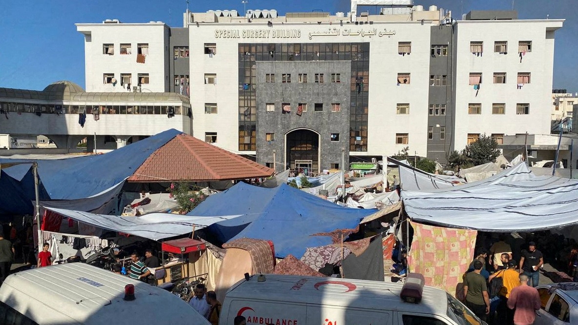 Makeshift tents cover the area in front of a multi-storey stone hospital building, two ambulances are in the foreground
