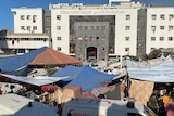 Makeshift tents cover the area in front of a multi-storey stone hospital building, two ambulances are in the foreground