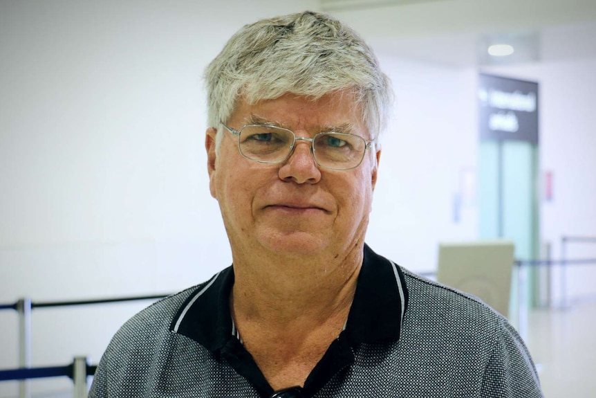 An older man with grey hair and glasses waits in an airport terminal