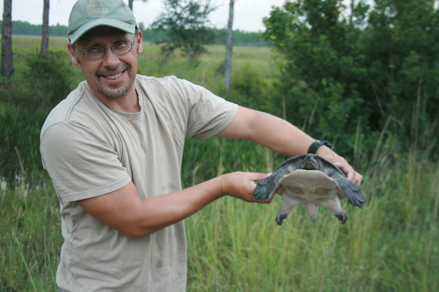 A smiling man, wears cap, glasses, stands in grassy area,  holds a turtle.