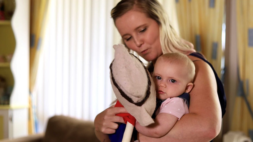 A blonde woman sits looking at her baby girl who is holding a doll