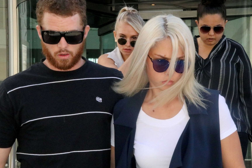 Close headshot of three women and a man all wearing sunglasses, looking down, outside court.