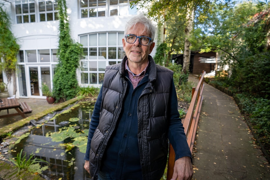 A grey-haired man with glasses dressed in a gilet stands in a garden.
