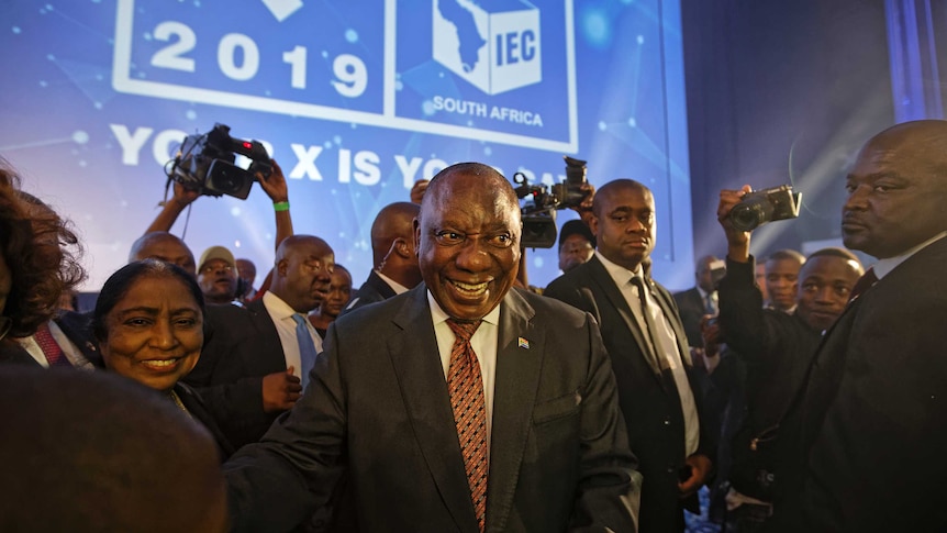 President Cyril Ramaphosa smiles boadly as he's surrounded by supporters, in font of a blue screen