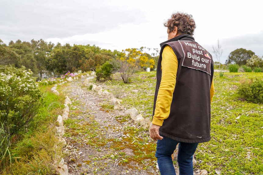 An woman wearing jeans and a black vest with the words 'heal our past, build our future' walks along a path bordered by flowers.