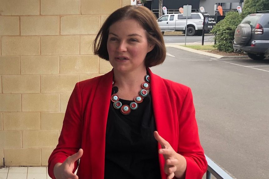 A short-haired woman wears a red jacked and a black top and a big necklace gestures as she speaks to the camera in a street.