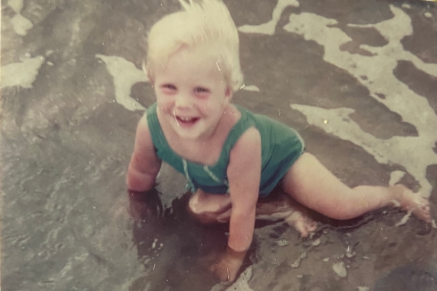 A toddler with white hair in a swimming suit wades in the water.