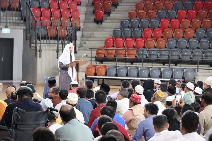 A man at a lectern speaking to a large group of people sitting on the ground, inside an indoor sports stadium.