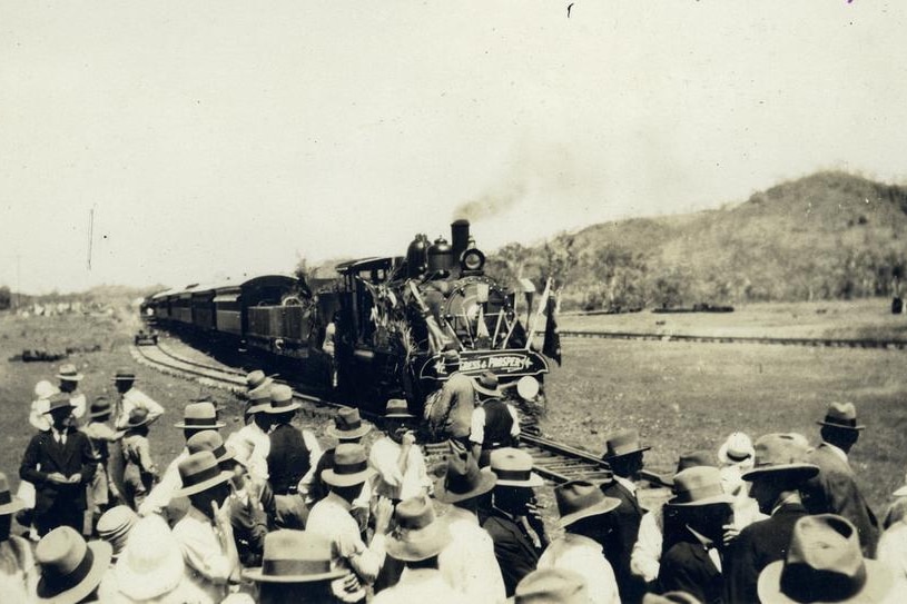 A black-and-white image of people in hats watching a steam train approach on an outback rail line.